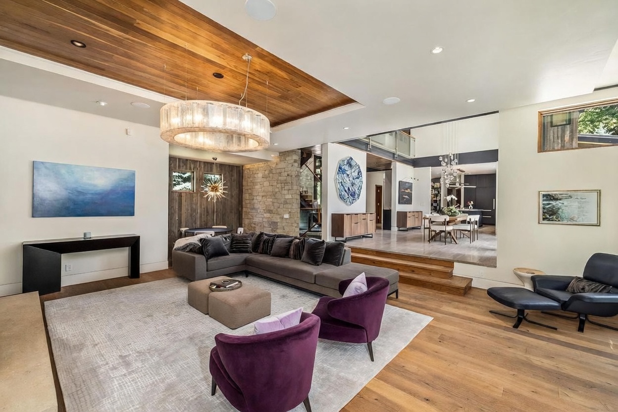 This expansive, open-concept living area features a sunken seating arrangement centered around a large gray sectional and two vibrant purple armchairs. The space is defined by a striking wood-paneled ceiling detail, a tall stone-clad wall, and a large circular chandelier that anchors the room. The perspective looks from the living area toward a raised dining space, showcasing a sophisticated, modern aesthetic with high-end finishes and a neutral color palette.
