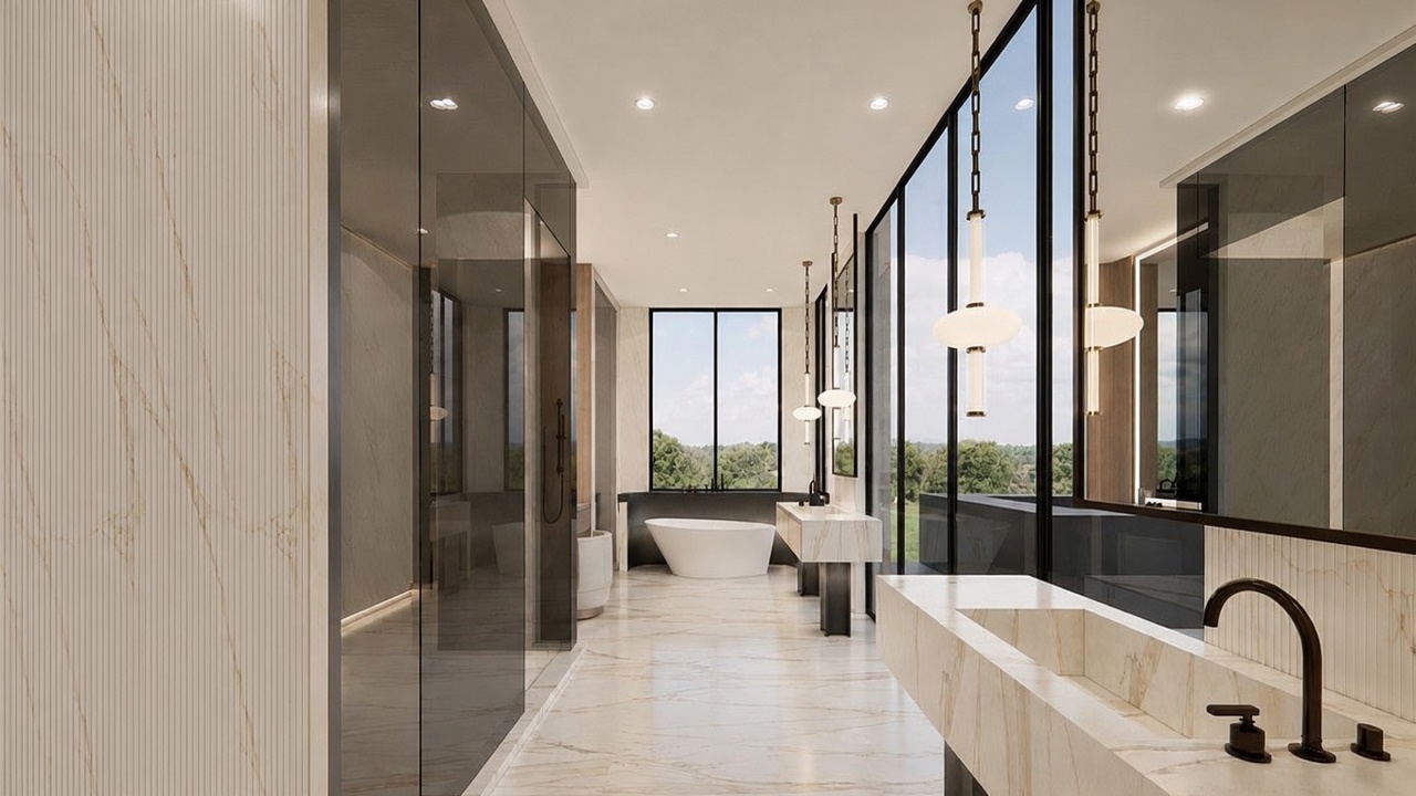 This luxurious primary bathroom features a sleek, modern design with a prominent stone-clad vanity and a freestanding soaking tub positioned in front of a large window. The space is defined by expansive glass partitions, polished stone flooring, and elegant pendant lighting that creates a sophisticated, spa-like atmosphere. The perspective looks down the length of the room, highlighting the clean lines and high-end finishes throughout.