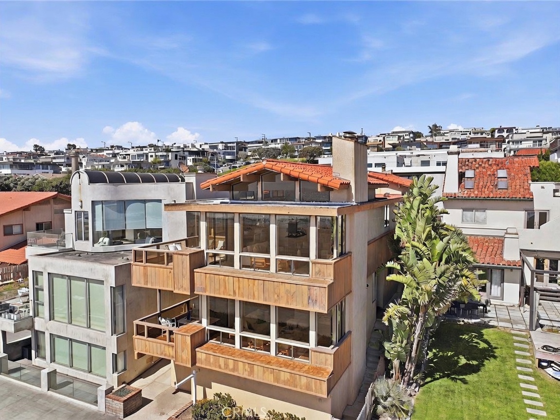 This elevated perspective captures a multi-story coastal residence featuring a distinctive wood-paneled facade and large windows that offer expansive views. The home includes stacked balconies and a classic terracotta-tiled roof, set within a dense neighborhood of similar hillside properties. A lush green lawn sits to the side, framed by a large bird of paradise plant, creating a bright and airy atmosphere.