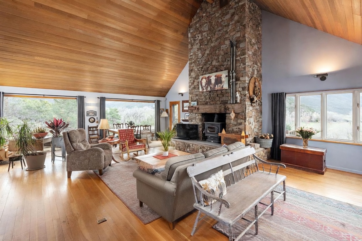 This spacious living room features a vaulted wood-paneled ceiling and a prominent stone-clad chimney breast that serves as the room's focal point. The space is furnished with a comfortable sofa, a rustic wooden bench, and a cozy armchair, all arranged around a central coffee table. Large windows provide expansive views of the surrounding landscape, filling the room with natural light and creating an inviting, mountain-inspired atmosphere.