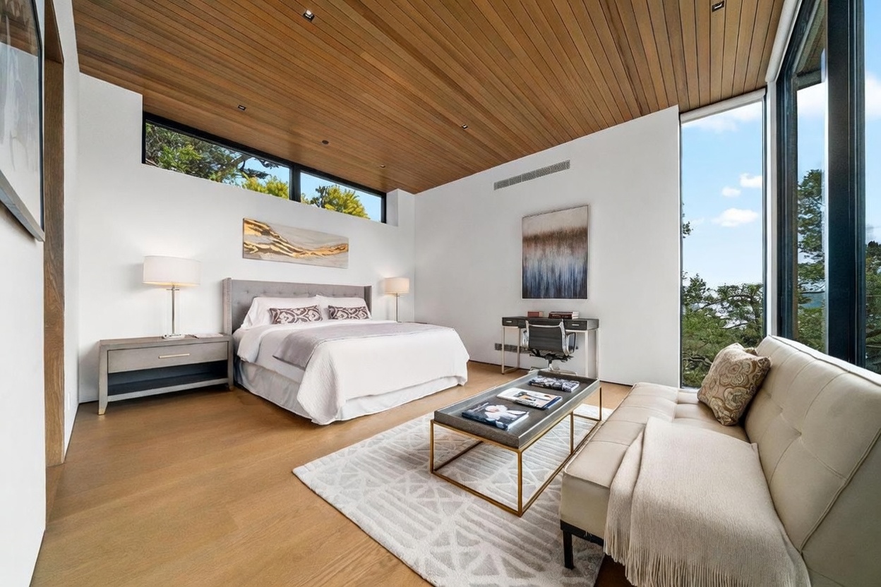 This spacious primary bedroom features a warm wood-paneled ceiling and light wood flooring, creating a serene and modern atmosphere. The room is furnished with a plush bed, a sleek desk area, and a comfortable seating arrangement, all complemented by large windows that provide expansive views of the surrounding trees. The minimalist aesthetic is enhanced by neutral tones and contemporary artwork, offering a sophisticated retreat.