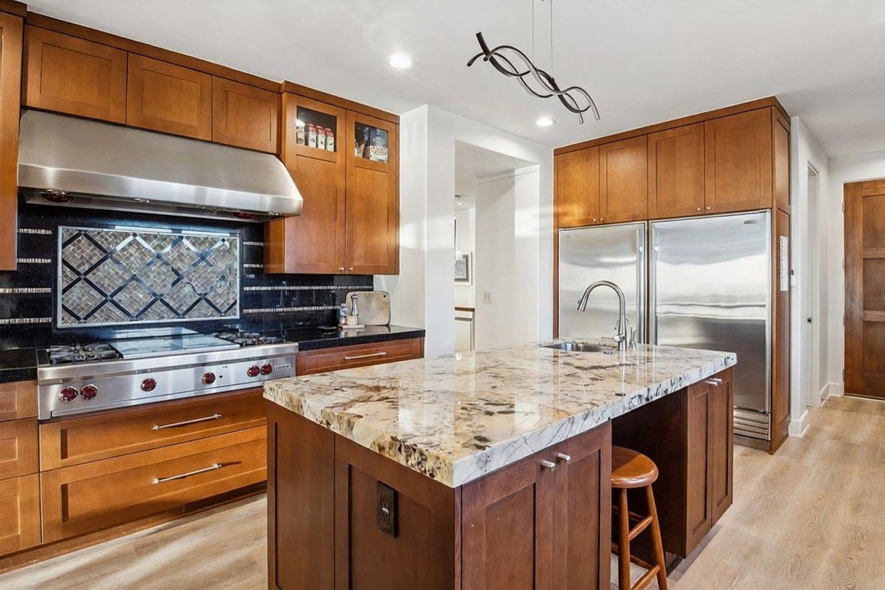 This kitchen features warm-toned wood cabinetry, a large central island with a stone countertop, and professional-grade stainless steel appliances. A decorative tile backsplash sits behind the gas range, while a modern light fixture hangs above the island. The space is finished with light-colored flooring, creating a bright and inviting atmosphere.