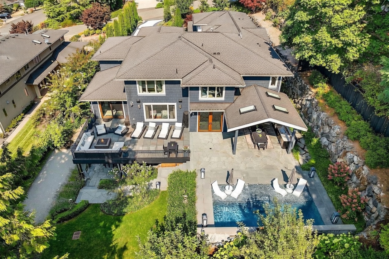 This high-angle aerial view captures a luxurious multi-story residence with a dark-toned shingle roof and blue-grey siding. The property features an expansive outdoor living area, including a raised deck with a fire pit, a covered patio with an outdoor kitchen, and a swimming pool with integrated lounge chairs. The backyard is beautifully landscaped with stone retaining walls, lush greenery, and a manicured lawn, creating a sophisticated and private retreat.