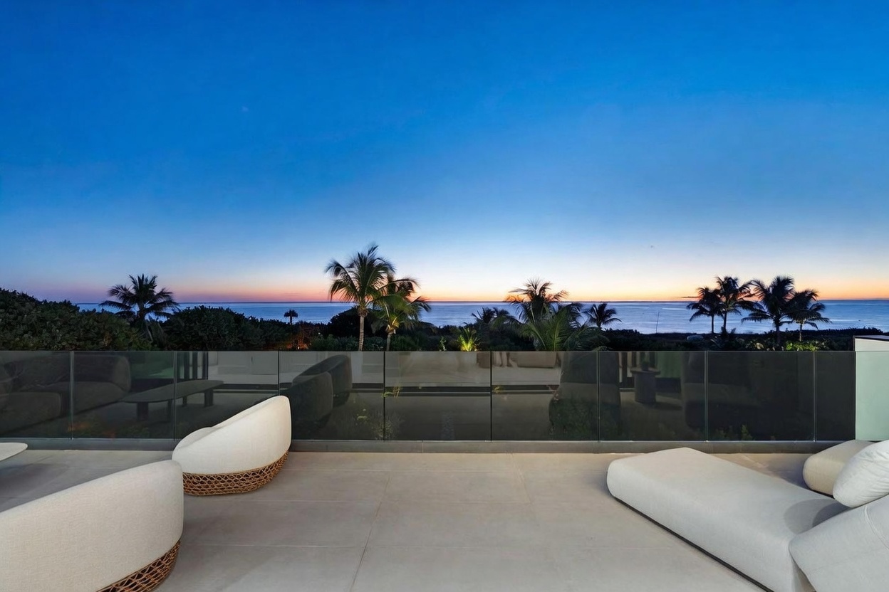 This expansive balcony offers a serene, elevated vantage point overlooking the ocean at twilight. The space is furnished with modern, low-profile lounge seating and features a sleek glass railing that ensures an unobstructed view of the horizon and palm trees. The neutral-toned flooring and minimalist furniture create a sophisticated, tranquil atmosphere perfect for relaxation.