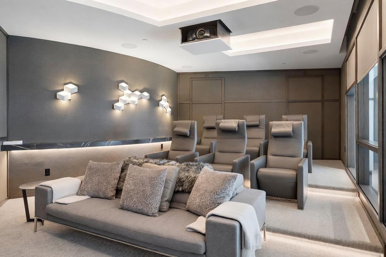 This sophisticated home theater features a tiered layout with a plush gray sofa in the foreground and three rows of leather reclining seats behind it. The room is accented by geometric wall sconces, a projector mounted on the ceiling, and a sleek, dark stone ledge with integrated lighting. The space is designed for comfort and entertainment, utilizing a neutral color palette and layered lighting to create an immersive cinematic atmosphere.