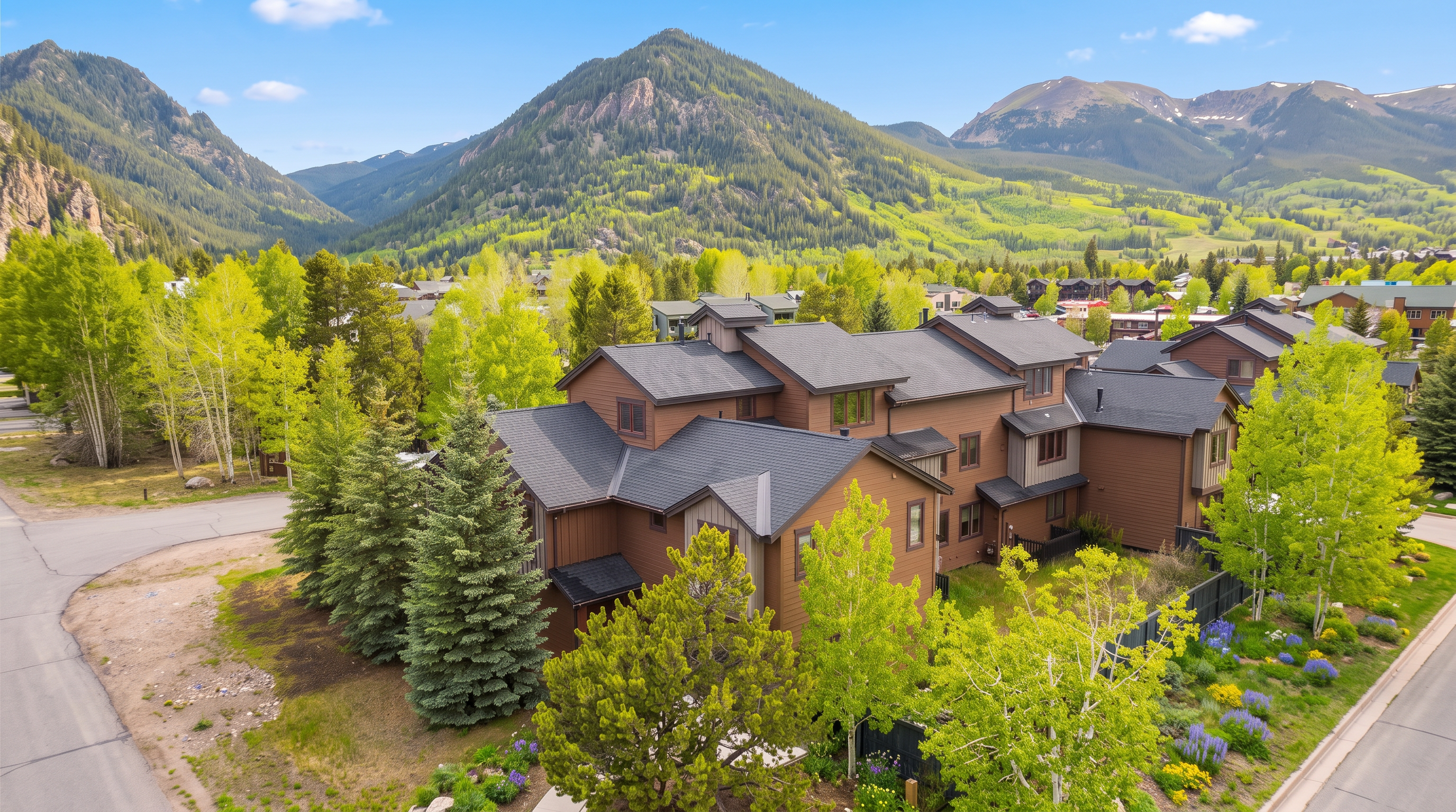 This elevated aerial perspective captures a multi-story mountain townhome complex nestled against a dramatic backdrop of rugged peaks at sunset. The architecture features a blend of brown horizontal siding and vertical accents, complemented by dark shingled roofs and warm, inviting interior lighting glowing through the windows. Surrounded by mature pine trees and aspen groves, the property offers a quintessential alpine aesthetic that emphasizes its serene, natural setting.