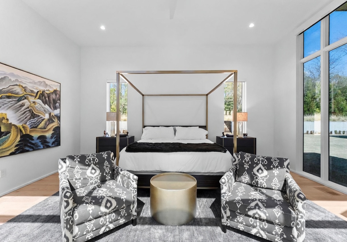 This spacious primary bedroom features a central four-poster bed with a metallic frame, flanked by two dark nightstands and matching lamps. In the foreground, two patterned armchairs face a central gold-toned drum table, all set atop a plush gray area rug. The room is brightened by large windows that offer a view of the outdoors, while a large landscape painting of the Great Wall of China serves as a striking focal point on the left wall.