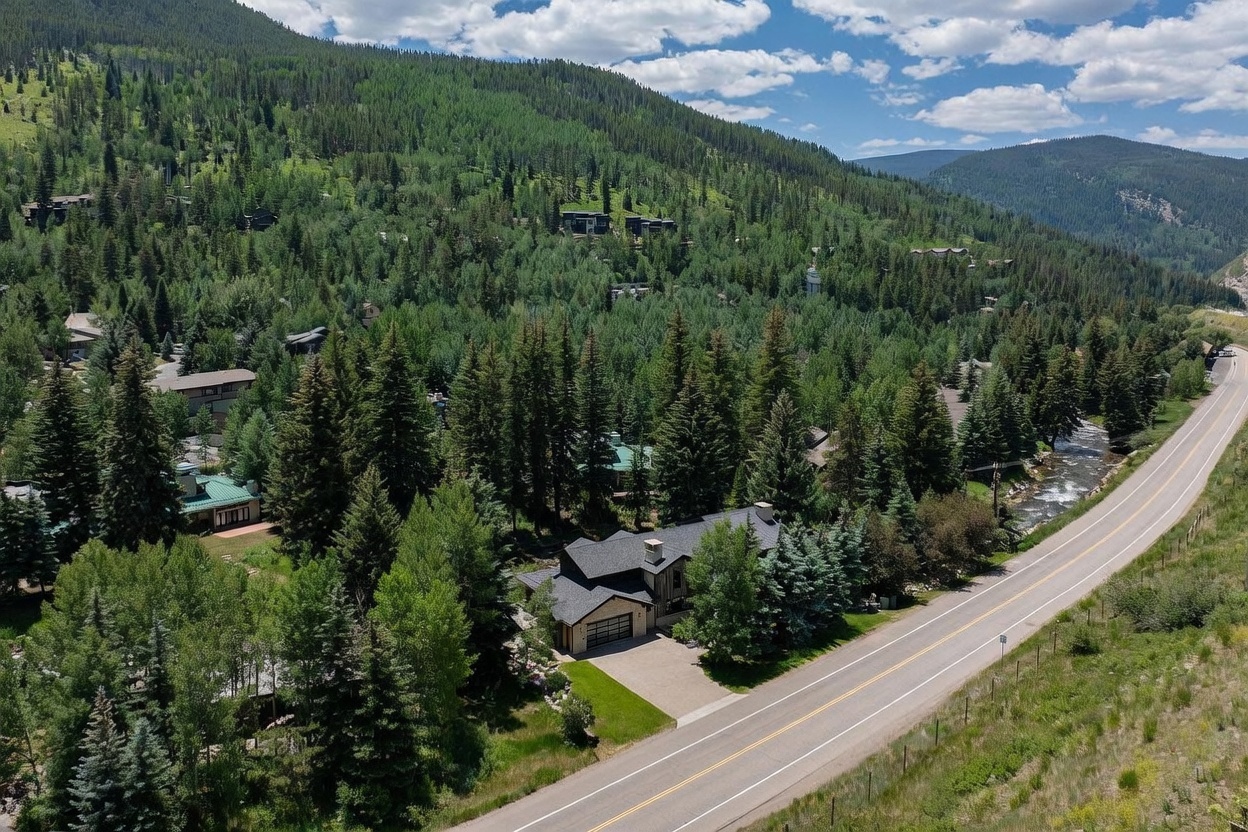 This high-angle aerial shot captures a multi-story mountain home nestled within a dense forest of pine and aspen trees. The property features a dark shingled roof, a paved driveway, and a garage, situated directly adjacent to a winding road and a flowing mountain stream. The surrounding landscape showcases a lush, mountainous terrain under a bright, partly cloudy sky, emphasizing the home's secluded and scenic location.