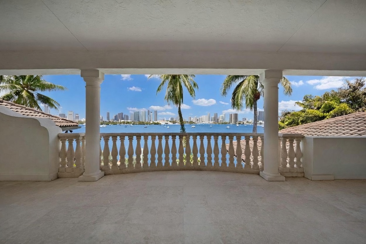 This covered balcony features a classic architectural style with white columns and a decorative stone balustrade. The open-air space offers a panoramic view of the bay and the distant city skyline, framed by palm trees. The neutral-toned stone flooring creates a seamless transition from the interior to the outdoor living area.