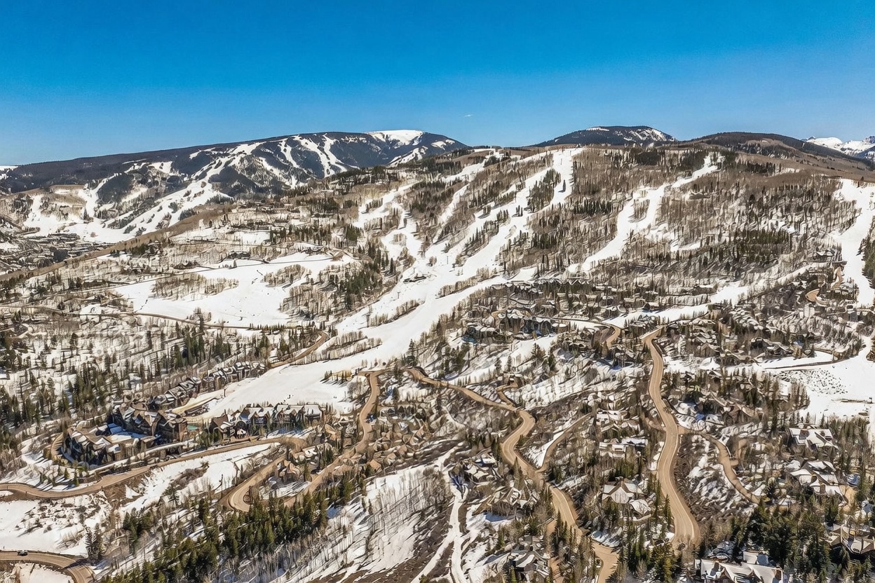 This expansive aerial view captures a sprawling mountain resort community nestled into a snowy, forested landscape. The perspective highlights the winding roads, clustered residential properties, and direct proximity to the groomed ski runs of the mountain slopes. The bright, clear blue sky contrasts with the snow-covered terrain, emphasizing the prime location for winter sports enthusiasts.
