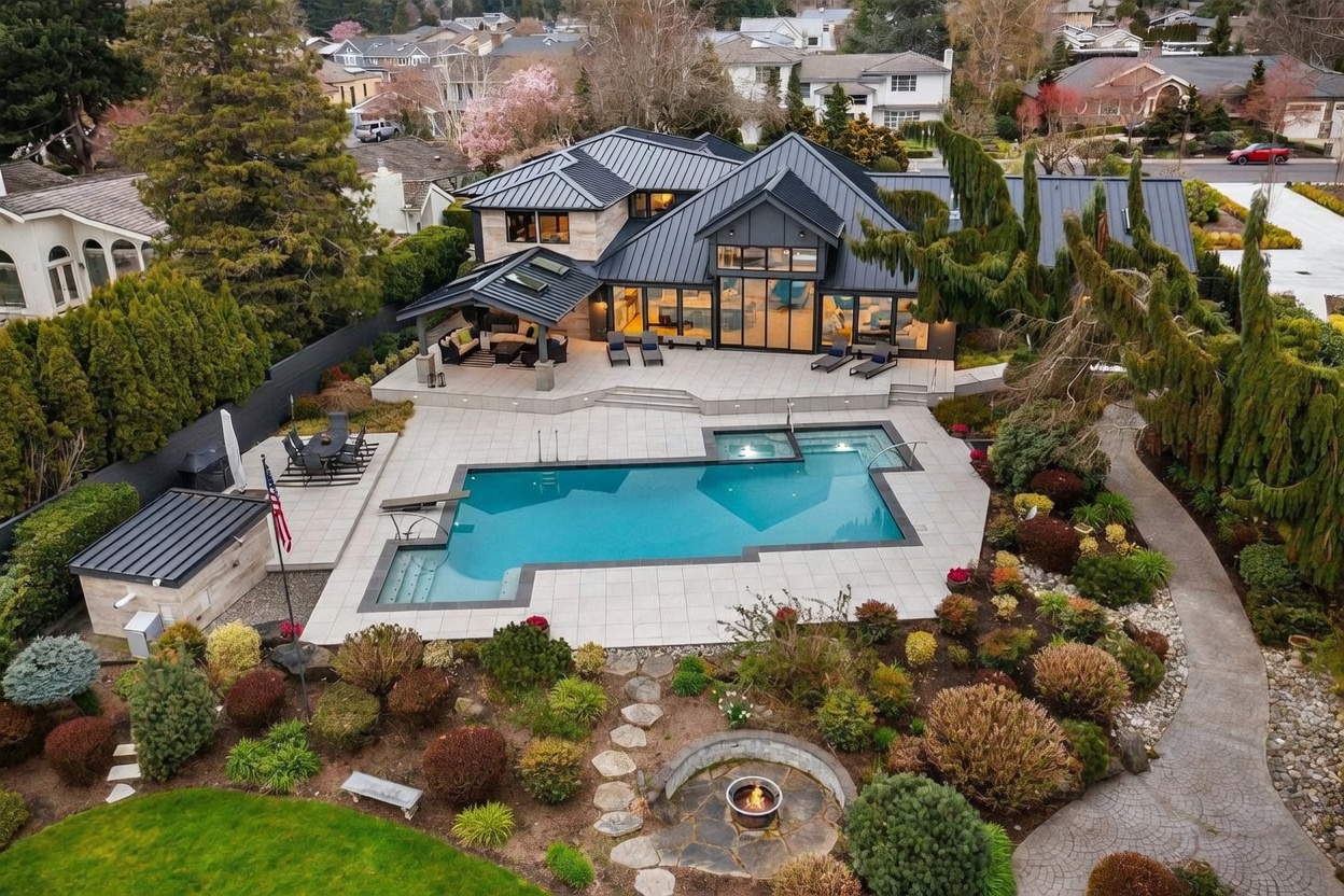 This high-angle aerial view showcases a luxurious modern residence with a striking dark metal roof and stone accents, seamlessly integrated into a beautifully landscaped backyard. The property features an expansive stone patio, a large swimming pool with an integrated spa, a covered outdoor living area, and a sunken fire pit, creating an ideal environment for high-end entertaining. The lush, manicured gardens and mature trees provide a sense of privacy and elegance to this sophisticated suburban estate.