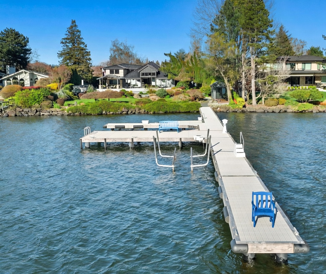 This eye-level shot captures a serene waterfront property from the perspective of a dock extending into the lake. The home features a modern architectural style with a dark roof and large windows, set against a lush, landscaped lawn filled with mature trees and shrubs. The scene conveys a peaceful, high-end lifestyle, emphasizing the seamless connection between the residence and the water.