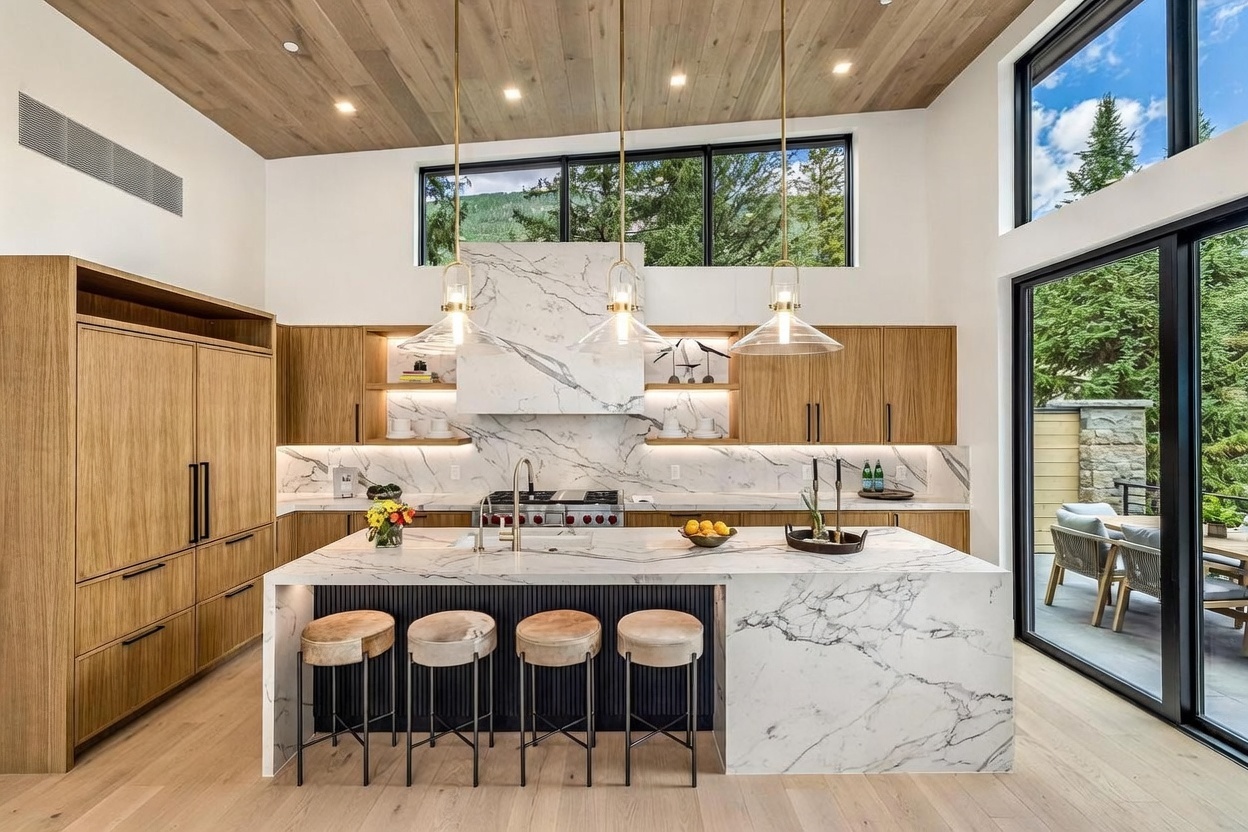 This modern kitchen features a large central island with a stone countertop and integrated seating, complemented by light wood cabinetry and a striking stone-clad backsplash. The space is illuminated by three glass pendant lights and benefits from natural light streaming through a wide window above the cooking area. A seamless transition to an outdoor patio is provided by large glass sliding doors, creating an airy and sophisticated atmosphere.