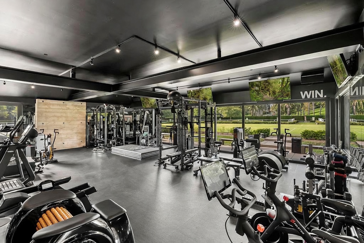 This is a spacious and well-equipped home gym featuring a variety of exercise machines, including treadmills, stationary bikes, and weightlifting equipment. The room has a modern aesthetic with a dark ceiling, sleek flooring, and large windows that offer views of the outdoors. The layout is designed for functionality and provides ample space for various workout activities.