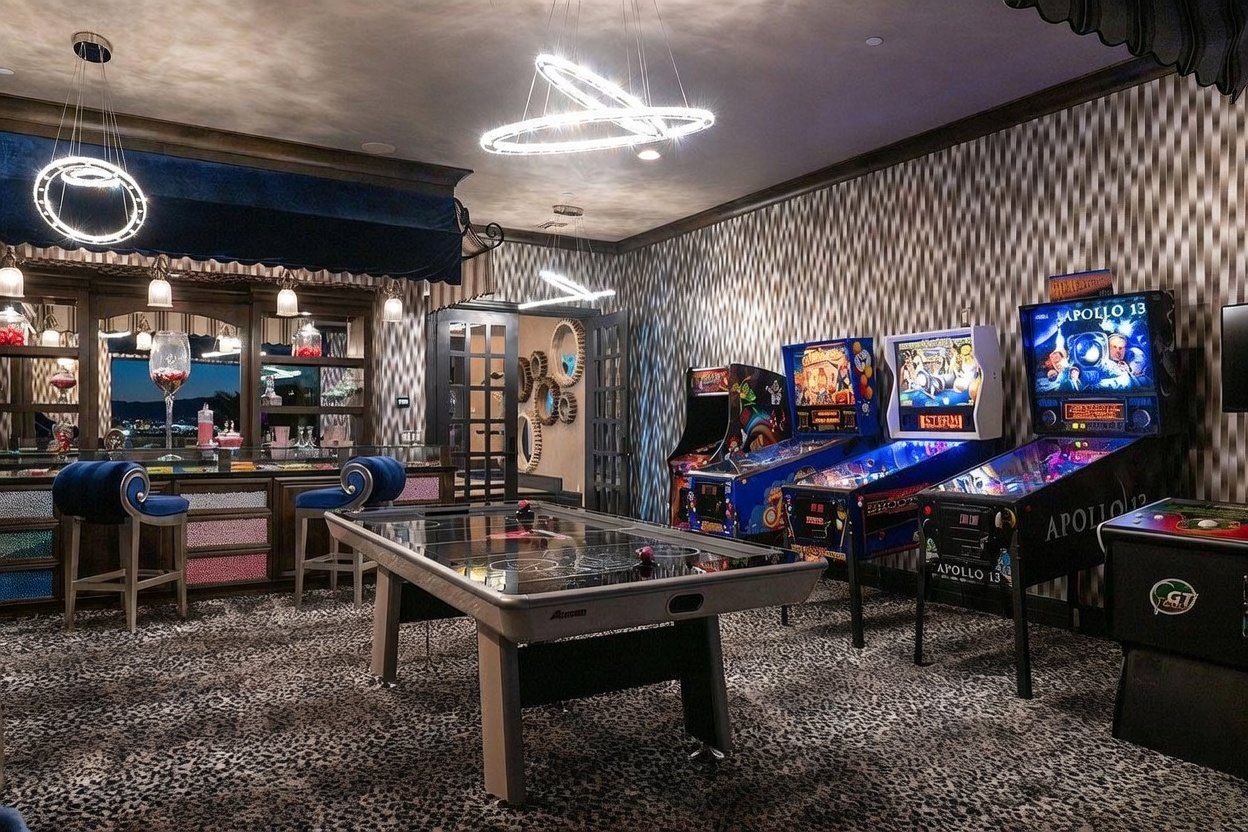 This vibrant game room features a central air hockey table set against a backdrop of patterned carpeting and a wall with a geometric wallpaper design. To the left, a built-in bar area with blue velvet stools and decorative lighting creates a social atmosphere, while the right side is lined with a row of classic pinball machines. The room is illuminated by modern, circular pendant light fixtures, contributing to a high-energy, entertainment-focused aesthetic.