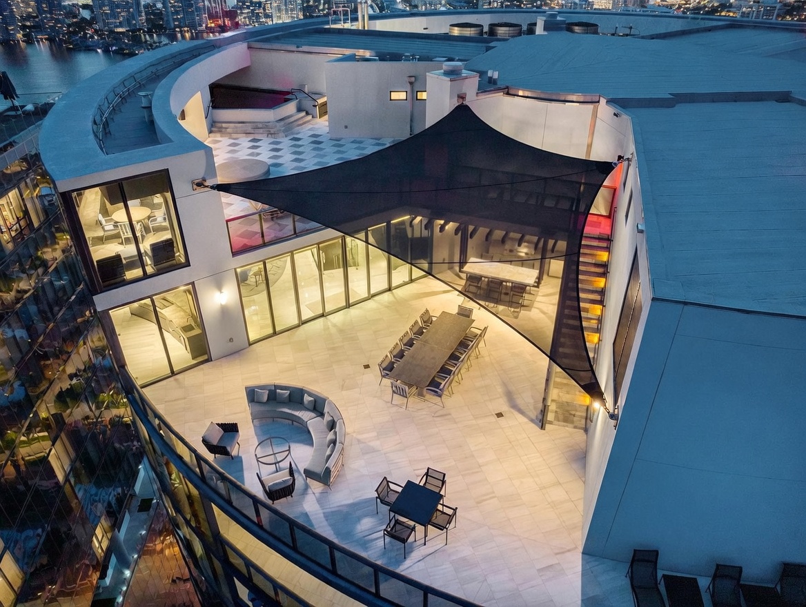 This high-angle aerial view captures a spacious, modern rooftop terrace at dusk, featuring a large dining area shaded by a geometric fabric canopy. The expansive deck is furnished with a curved sectional sofa, a separate dining set, and additional lounge seating, all set against a backdrop of city lights and a waterfront. The sleek architecture includes large glass walls that provide a seamless transition between the indoor living spaces and the outdoor entertainment area.