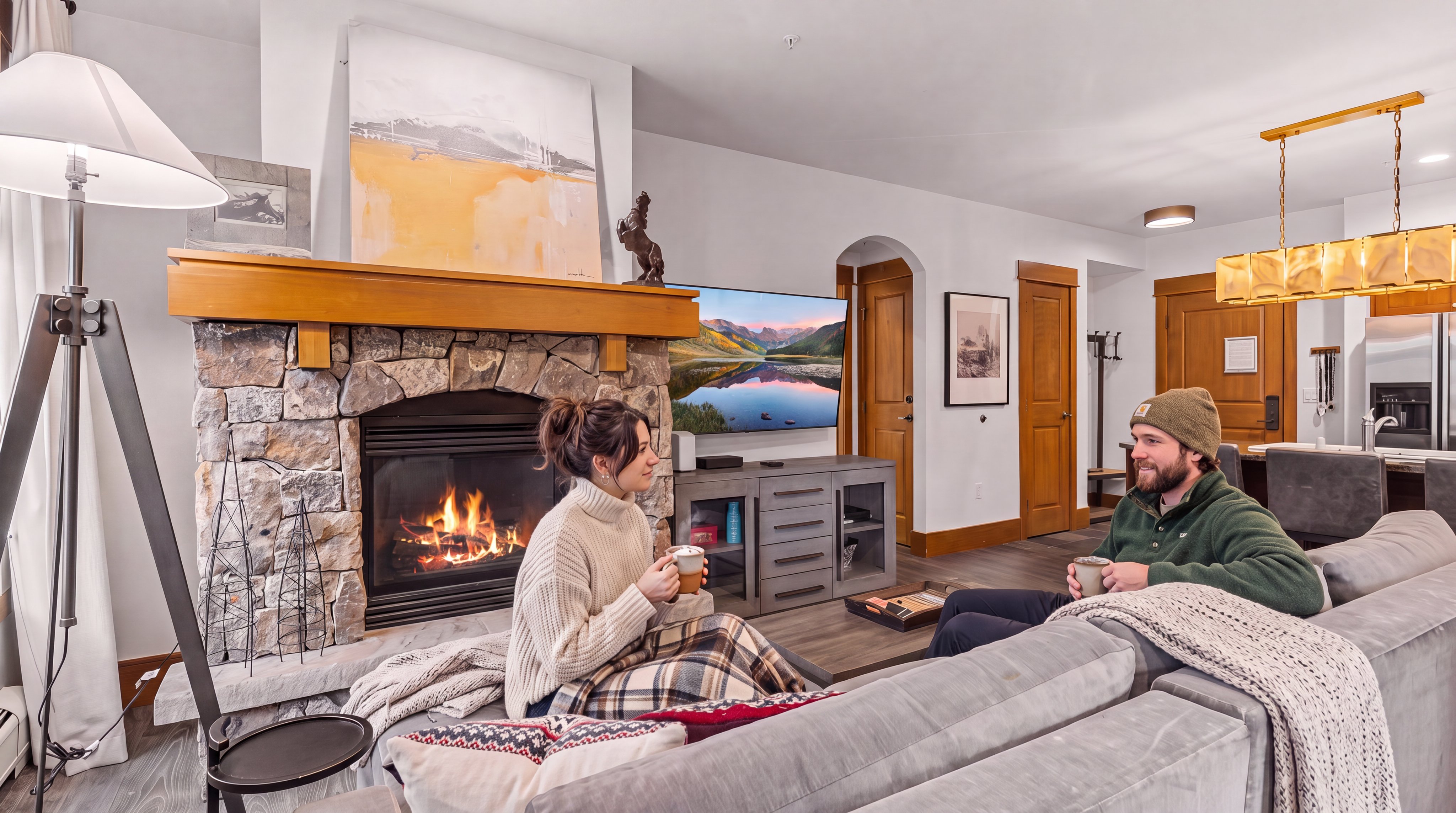 This inviting living room features a prominent stone-clad fireplace with a wooden mantel, serving as the focal point of the space. A large gray sectional sofa provides comfortable seating, while a television is mounted on the wall adjacent to the fireplace above a gray media console. The open-concept layout flows into a dining area with a modern light fixture, creating a cohesive and warm atmosphere suitable for relaxation or entertaining.