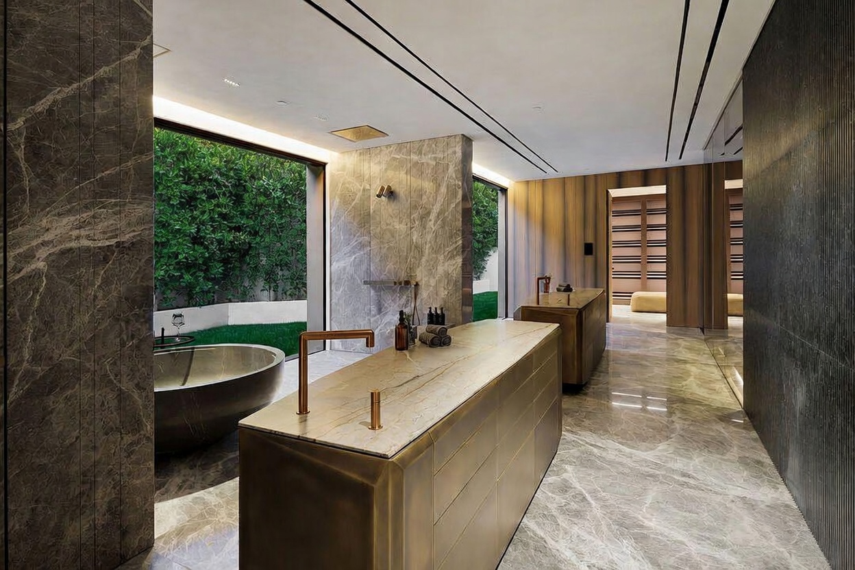 This luxurious primary bathroom features a sophisticated blend of marble surfaces, a sleek metallic vanity island, and a deep soaking tub positioned next to a floor-to-ceiling glass wall overlooking a private garden. The space exudes a high-end, spa-like atmosphere with its minimalist design, warm ambient lighting, and seamless transition between indoor and outdoor living. The perspective captures the expansive layout, highlighting the premium materials and the serene, nature-integrated aesthetic.