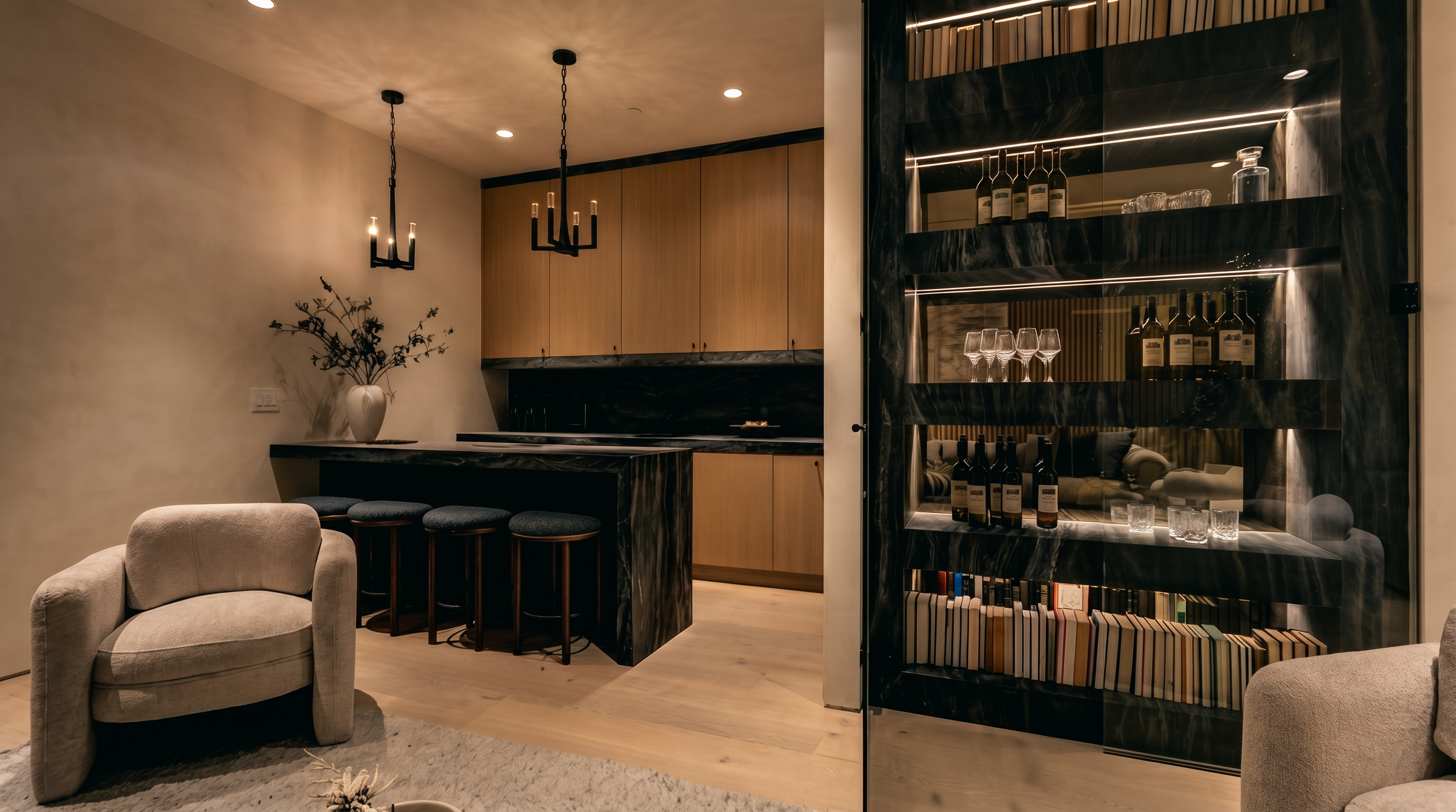 This sophisticated interior space features a modern bar area with a dark stone-clad island and matching countertop, complemented by light wood cabinetry. To the right, a glass-enclosed display wall showcases wine bottles and books, illuminated by integrated lighting. The room is furnished with a plush armchair in the foreground, creating a luxurious and inviting atmosphere.