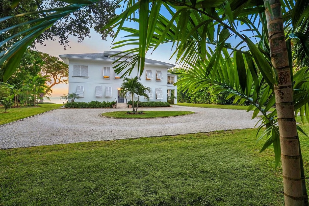 This image captures the front exterior of a stately, multi-story white home featuring classic architectural details and shuttered windows. A large, circular gravel driveway centers around a manicured lawn with a small palm tree, framed by lush tropical foliage and mature trees in the foreground. The perspective is taken from a low angle, looking through palm fronds toward the residence, creating a sense of privacy and grandeur.