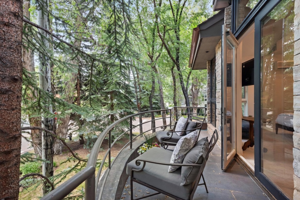 This elevated balcony features a curved metal railing and stone flooring, providing a serene outdoor seating area nestled among mature trees. Two comfortable armchairs with patterned cushions are arranged to overlook the wooded surroundings, creating a peaceful retreat. The space connects directly to the interior through a glass door, blending indoor and outdoor living.