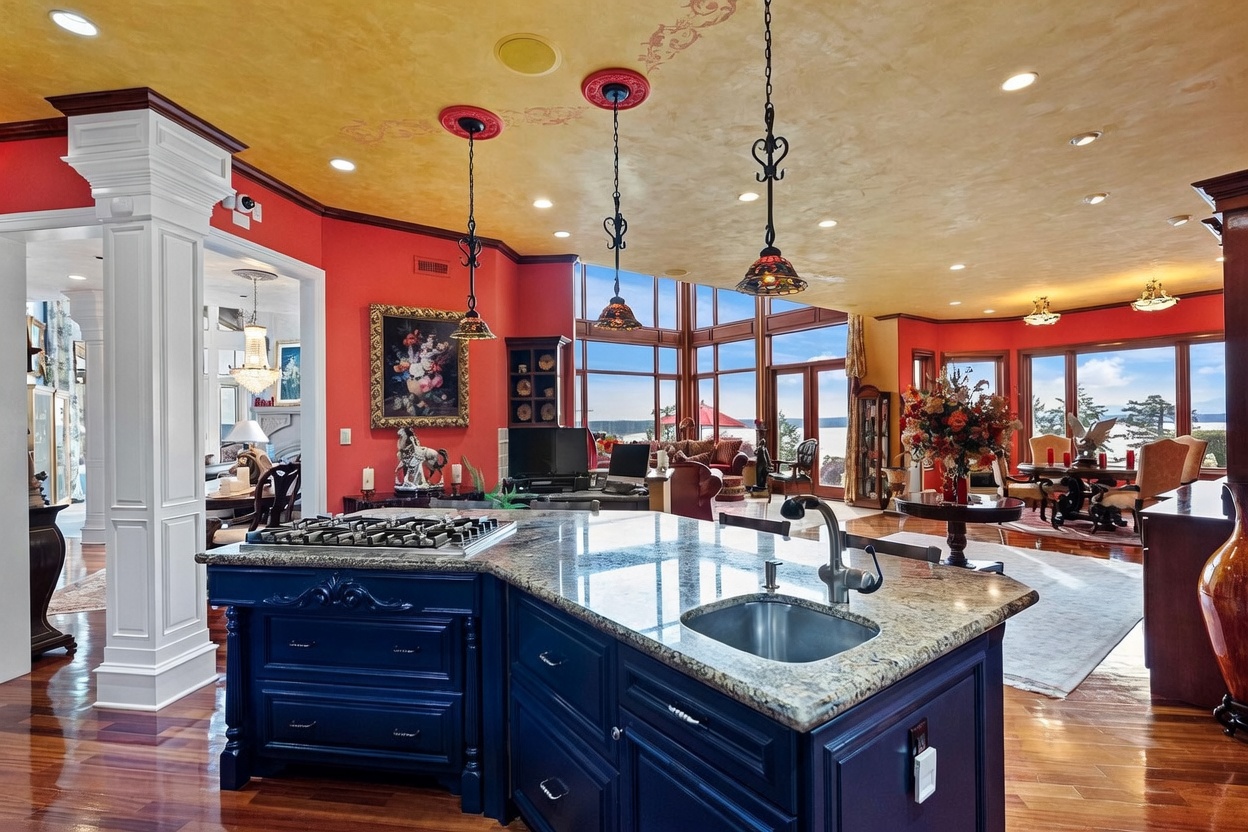 This spacious open-concept kitchen features a large, deep blue island with a stone countertop and an integrated sink, serving as the focal point of the room. The space is characterized by vibrant red walls, a textured yellow ceiling, and rich wood flooring that extends into the adjacent living and dining areas. Large windows provide expansive views of the outdoors, while decorative pendant lights hang above the island, creating a warm and inviting atmosphere.