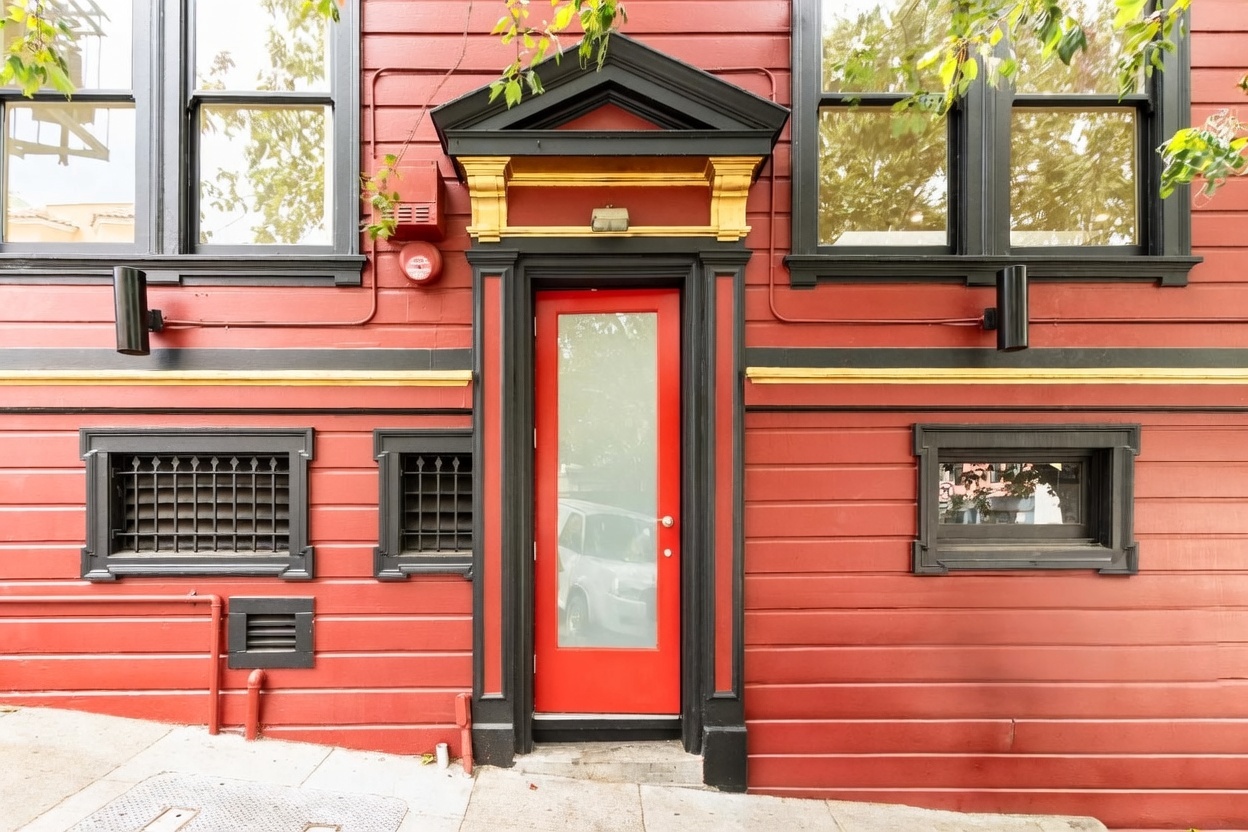 This striking entryway features a vibrant red door with a frosted glass panel, framed by bold black trim and a decorative pediment with gold accents. The exterior wall is clad in horizontal red siding, complemented by black-framed windows and symmetrical wall-mounted light fixtures. The composition is captured from a straight-on perspective, highlighting the architectural contrast and the clean, urban aesthetic of the building's facade.