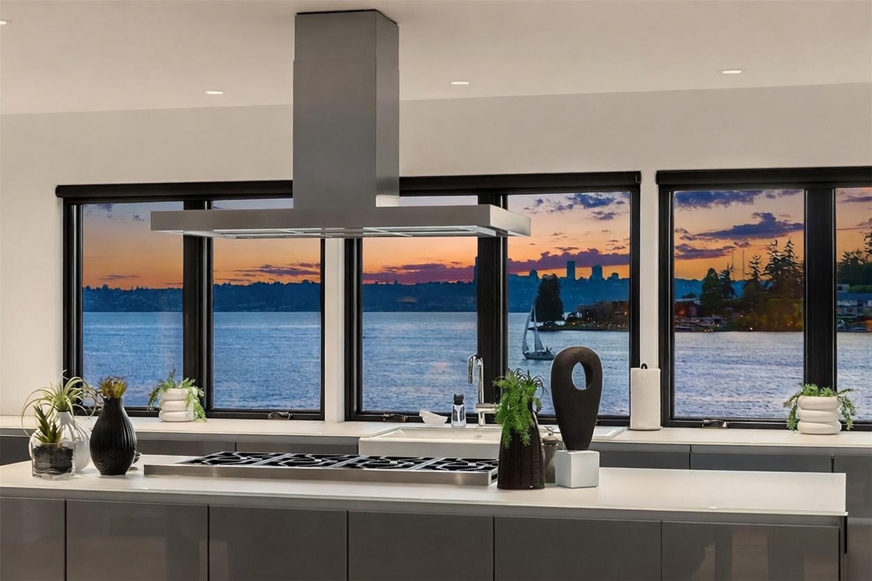 This modern kitchen features a sleek, dark-toned island with a built-in cooktop and a stainless steel range hood. Large windows behind the counter provide a stunning, unobstructed view of a lake and a distant city skyline at sunset, creating a dramatic and serene atmosphere. The space is accented with minimalist decor, including small potted plants and a sculptural vase, emphasizing a clean and sophisticated aesthetic.