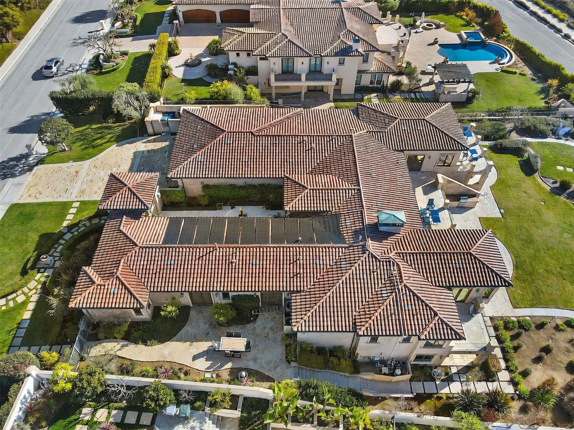 This high-angle aerial perspective captures a sprawling Mediterranean-style estate with a complex, multi-gabled clay tile roof. The property features a large central courtyard, a solar panel array integrated into the roofline, and extensive stone-paved patio areas surrounding the residence. Lush landscaping, manicured lawns, and a neighboring property with a swimming pool complete the expansive suburban setting.
