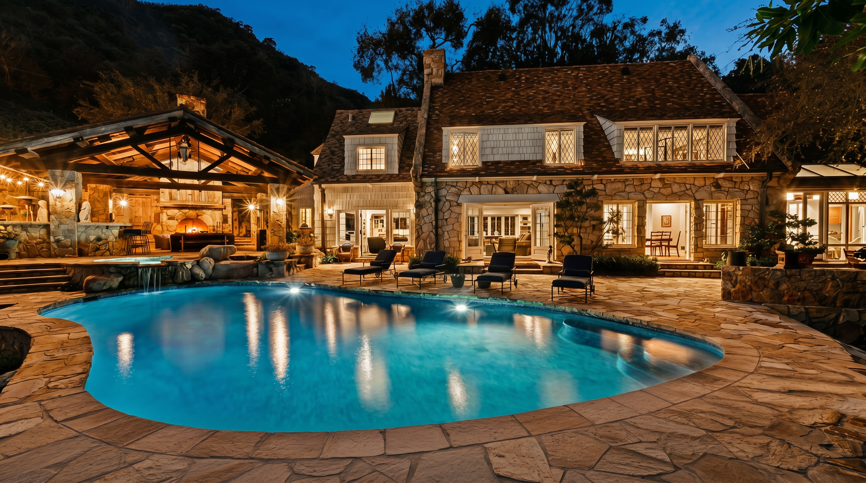 This stunning evening shot captures a luxurious stone-clad residence featuring a sprawling, illuminated swimming pool as the centerpiece. To the left, a rustic, open-air pavilion with a grand stone fireplace provides an inviting outdoor living space, while the main house showcases classic architectural details and warm interior lighting. The scene exudes an atmosphere of sophisticated relaxation and high-end outdoor entertaining.