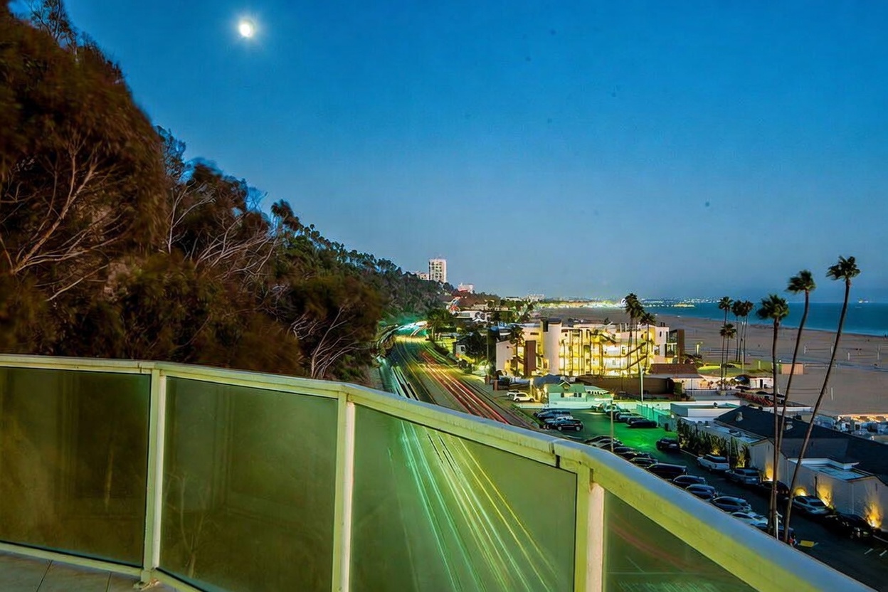 This image captures a stunning evening view from a balcony overlooking a coastal highway and the Pacific Ocean. The foreground features a modern glass-paneled railing, while the mid-ground shows the illuminated curves of the Pacific Coast Highway with light trails from passing cars. In the distance, the beach and the lights of the coastal town create a serene and picturesque atmosphere under a bright moon.