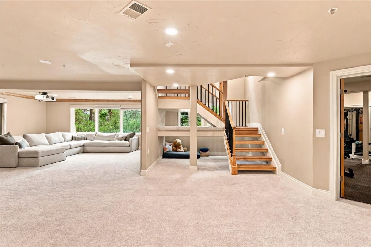 This spacious, finished basement features a large, neutral-toned sectional sofa arranged for comfortable lounging, complemented by a projector mounted on the ceiling. A unique architectural feature includes a cozy, built-in nook tucked beneath the wooden staircase, which is currently used as a pet area. The room is carpeted throughout and leads into a separate gym space visible through a doorway on the right, creating a versatile and inviting recreational area.
