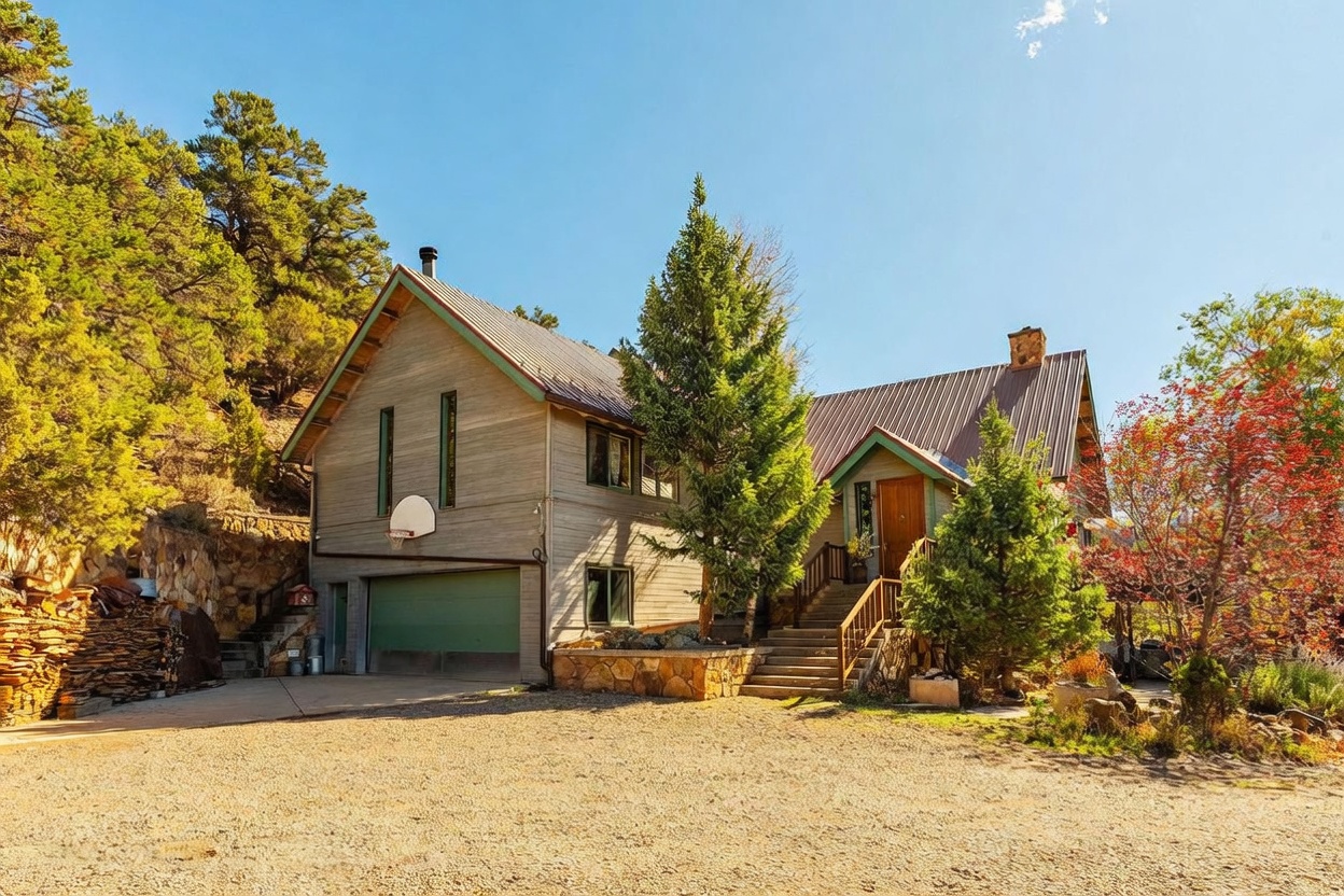 This mountain-style home features a multi-level design with light-toned wood siding and a metal roof, set against a backdrop of mature pine trees and a rocky hillside. The front exterior includes a prominent garage with a basketball hoop, a stone-accented foundation, and a welcoming staircase leading to the main entrance. The gravel driveway and surrounding natural landscaping create a rustic, secluded atmosphere typical of a mountain retreat.