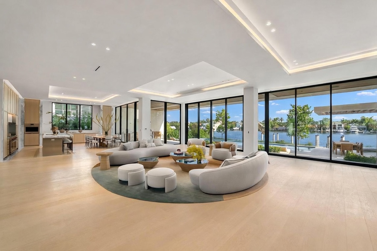 This expansive, open-concept living area features light wood flooring and a neutral color palette, centered around a circular rug with plush, curved sofas and modern coffee tables. Large, floor-to-ceiling glass walls provide an unobstructed view of the waterfront and outdoor patio, seamlessly blending indoor and outdoor living. The space is illuminated by recessed lighting and a sophisticated tray ceiling design, creating an airy and luxurious atmosphere.