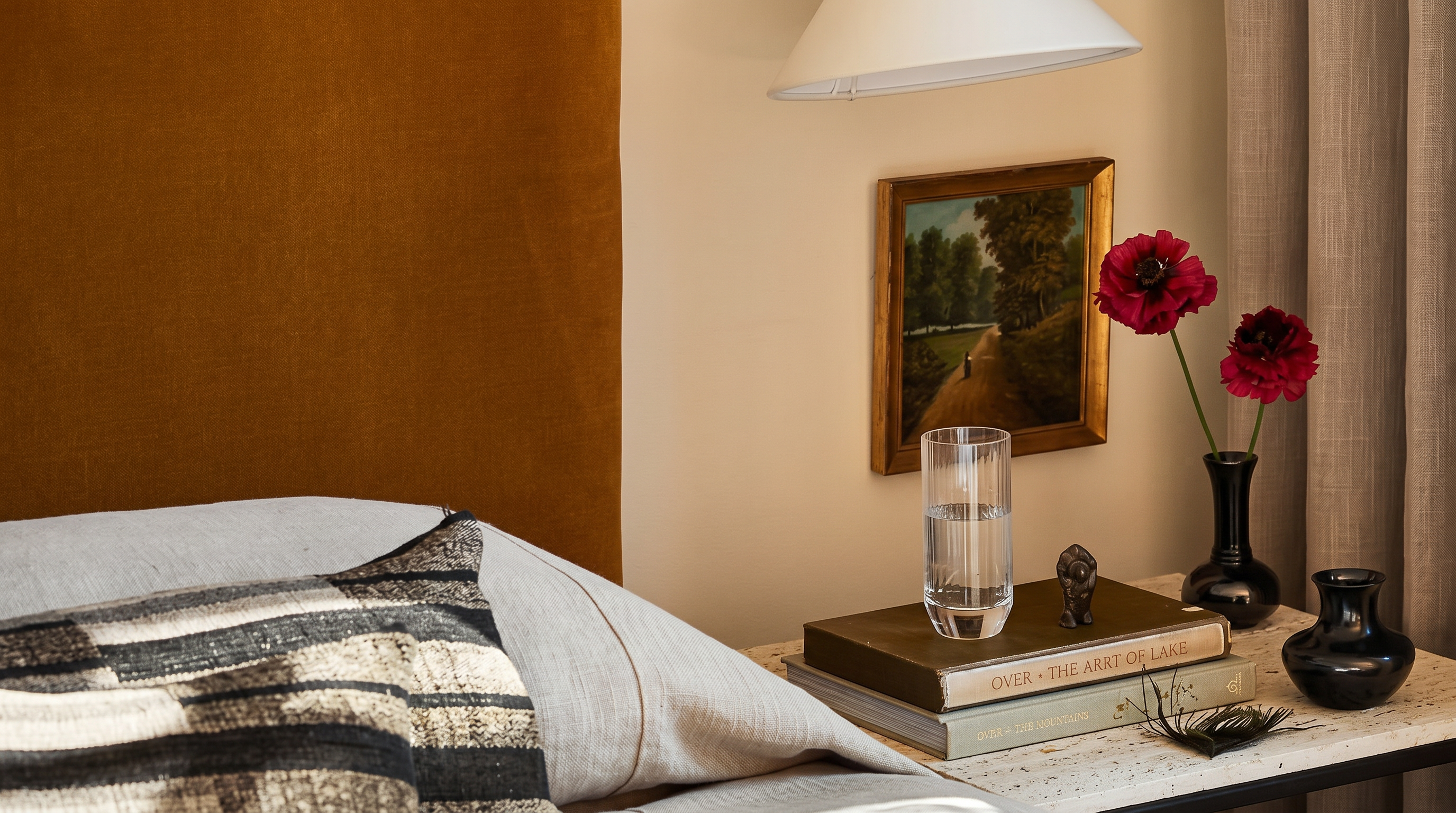 This cozy bedroom vignette features a plush, mustard-colored velvet headboard paired with neutral, textured bedding and a striped accent pillow. A minimalist white cone wall sconce provides warm lighting above a rustic wooden nightstand, which is adorned with a glass of water, a stack of books, and a small vase of deep red flowers. The scene is framed by soft, floor-length curtains, creating an inviting and sophisticated atmosphere.