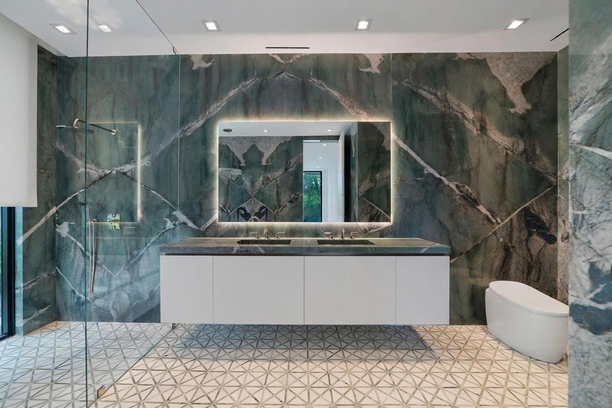 This luxurious primary bathroom features a striking wall clad in large-format green stone slabs with dramatic veining, serving as a bold backdrop for the floating double vanity. The space includes a modern white toilet, a glass-enclosed shower area, and a geometric patterned tile floor that adds visual texture. A large, rectangular mirror with integrated perimeter lighting enhances the sophisticated and contemporary aesthetic of the room.