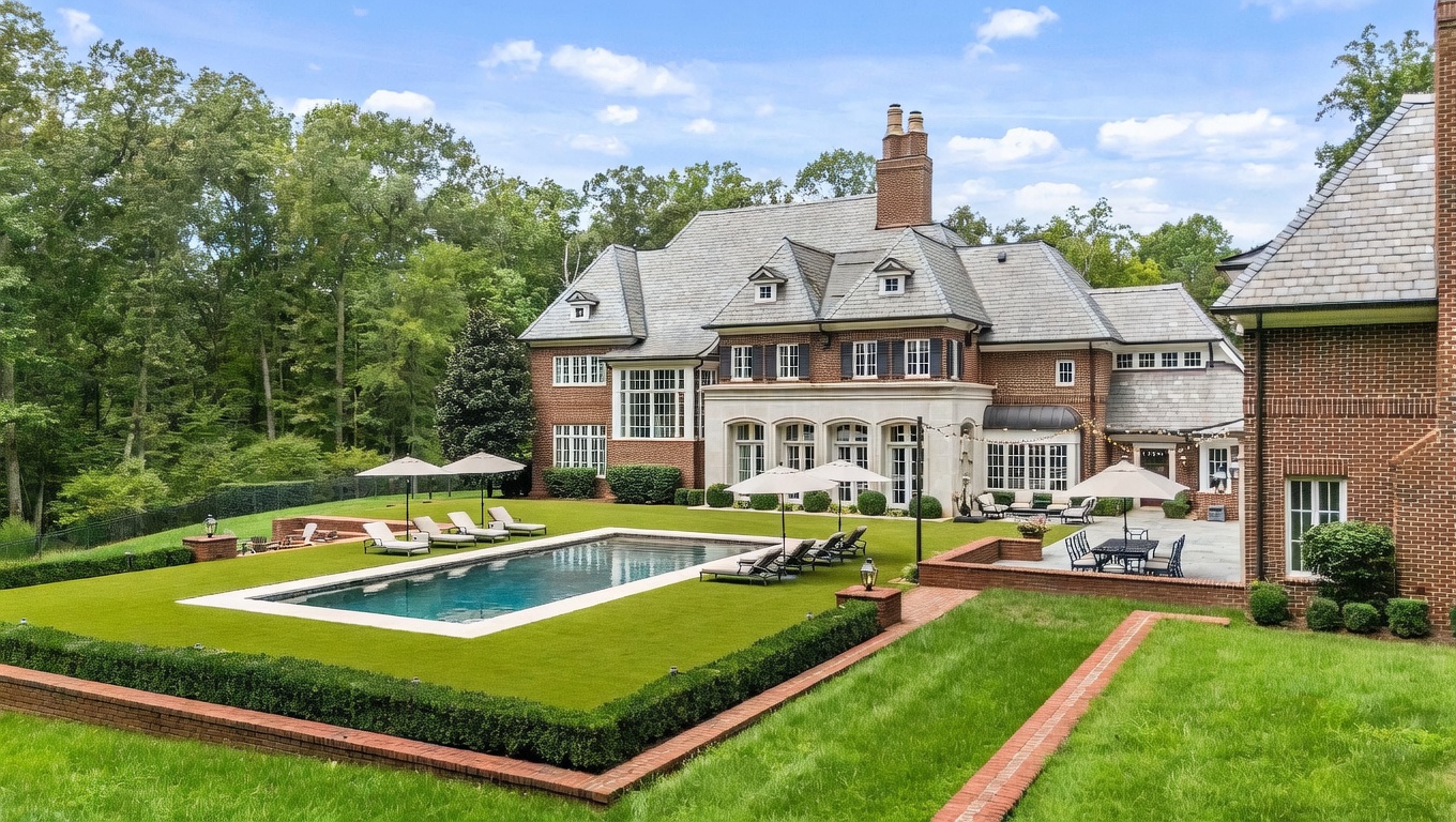 This expansive rear view of a grand brick estate showcases a sophisticated architectural design with multiple gables, dormer windows, and a slate roof. The property features a meticulously landscaped backyard with a rectangular swimming pool, a stone-paved patio area, and lush green lawns bordered by manicured hedges. The scene is captured from an elevated perspective, emphasizing the seamless transition between the elegant home and its private, wooded outdoor living space.