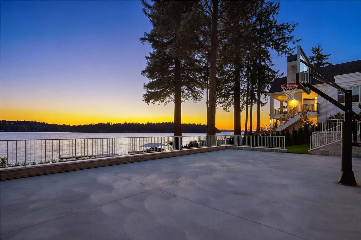This expansive concrete patio overlooks a serene lake at sunset, featuring a basketball hoop and a white metal railing that frames the water view. The scene includes mature trees and a glimpse of the home's multi-level exterior, which features a staircase leading to an upper balcony. The warm, golden light of the setting sun creates a peaceful and inviting atmosphere for outdoor recreation.