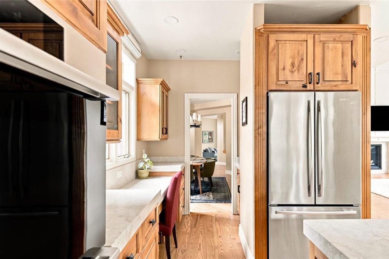 This kitchen features warm-toned wood cabinetry paired with light-colored stone countertops and a built-in desk area. A stainless steel refrigerator is integrated into the cabinetry, and the space flows through a doorway into an adjacent dining area. The perspective is from the kitchen looking toward the dining room, highlighting the open layout and hardwood flooring throughout.