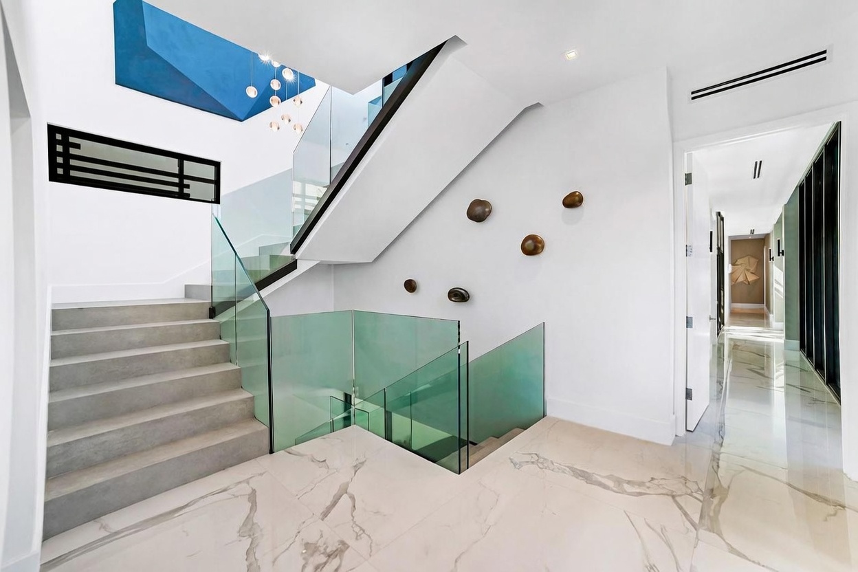 This modern entryway features a striking staircase with glass railings and light-toned steps, leading up toward a vibrant blue ceiling accent. The space is characterized by high-contrast design, including a geometric black wall feature and abstract circular wall art. Polished light-colored stone flooring with subtle veining extends into a bright, minimalist hallway, creating an open and sophisticated atmosphere.