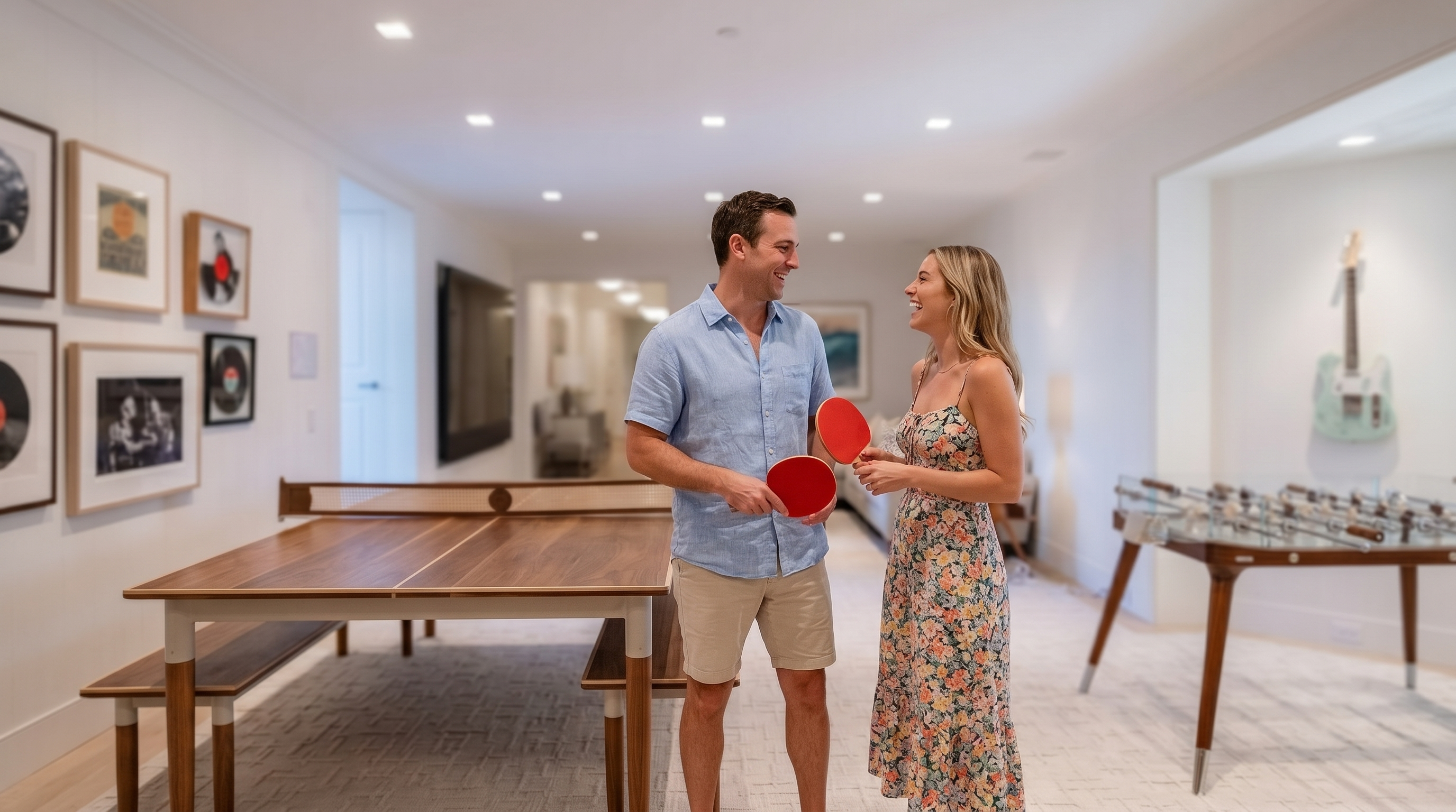 This spacious, modern basement recreation area features a high-end wooden ping pong table and a sleek glass-topped foosball table. The room transitions into a comfortable lounge area with a sectional sofa and a wall-mounted television, while a decorative electric guitar is mounted on the wall. The space is finished with neutral carpeting and recessed lighting, creating a bright and inviting atmosphere for entertainment.