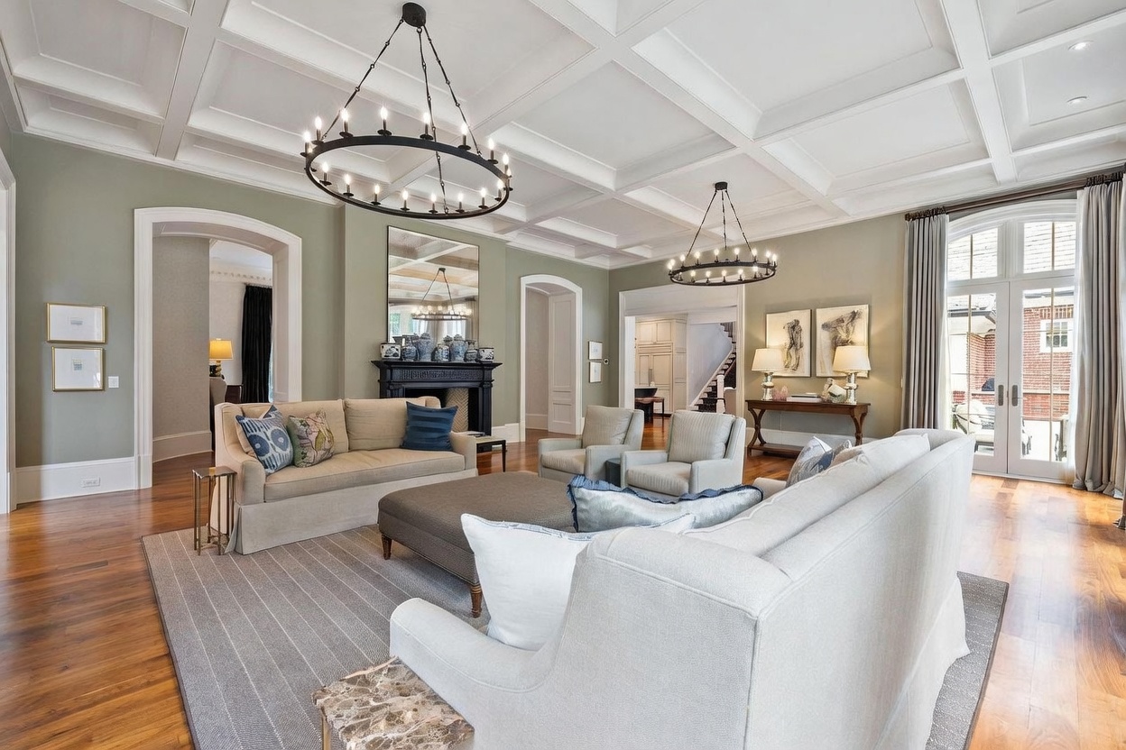 This elegant living room features a coffered ceiling with two large, circular chandeliers and light green walls. The space is anchored by a dark fireplace surround with a mirror above it, flanked by two arched doorways leading to other areas of the home. A neutral-toned sofa and armchairs are arranged on a large area rug, creating a sophisticated and inviting atmosphere.