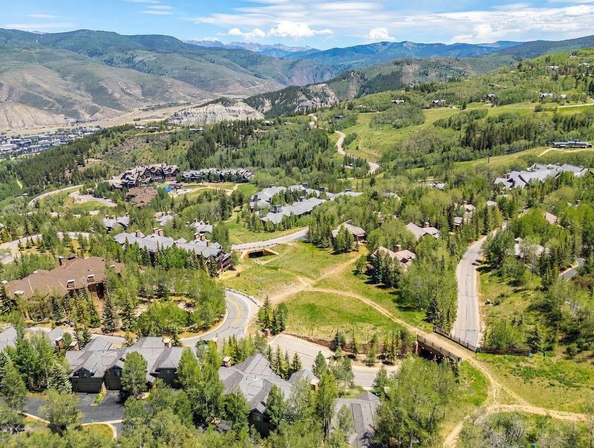 This high-angle aerial view captures a sprawling mountain community nestled within a lush, forested landscape. The scene features winding paved roads, clusters of multi-story residential buildings with dark shingled roofs, and expansive green spaces interspersed with mature trees. In the background, majestic mountain ranges stretch across the horizon under a bright, partly cloudy sky, emphasizing the serene and secluded nature of the location.