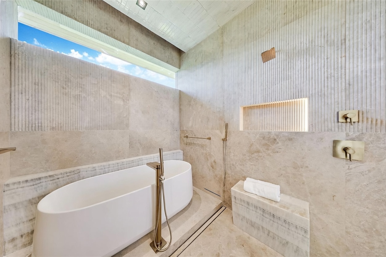 This luxurious, spa-inspired bathroom features a freestanding white soaking tub paired with a sleek, metallic floor-mounted faucet. The space is defined by textured, vertical-slat wall detailing and large-format stone tiles, complemented by a built-in stone bench and a recessed wall niche. A long, horizontal window above the tub allows natural light to flood the room, creating an airy and sophisticated atmosphere.