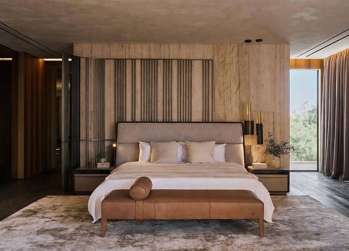 This luxurious primary bedroom features a sophisticated, neutral color palette with textured stone wall paneling and a sleek, modern headboard. A leather bench sits at the foot of the bed, which is dressed in crisp white linens, while floor-to-ceiling curtains frame a bright window view. The room exudes a high-end, serene atmosphere with minimalist decor and warm, ambient lighting.