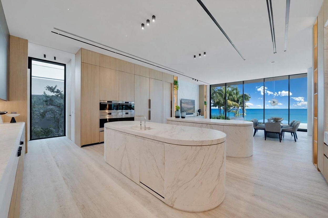 This modern, open-concept kitchen features dual curved islands with light-toned stone surfaces and integrated cabinetry. The space is defined by light wood cabinetry, recessed lighting, and expansive glass walls that provide an unobstructed view of the ocean. The minimalist design creates a seamless transition between the high-end culinary area and the adjacent dining space.