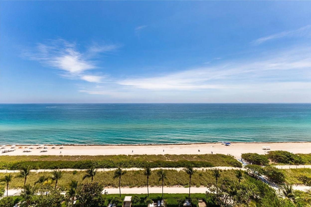 This elevated perspective captures a stunning, expansive view of the ocean horizon under a clear blue sky. In the foreground, a lush green landscape with palm trees and a sandy beach path leads directly to the shoreline, where gentle waves meet the sand. The scene conveys a serene, coastal lifestyle, highlighting the property's prime proximity to the beach.