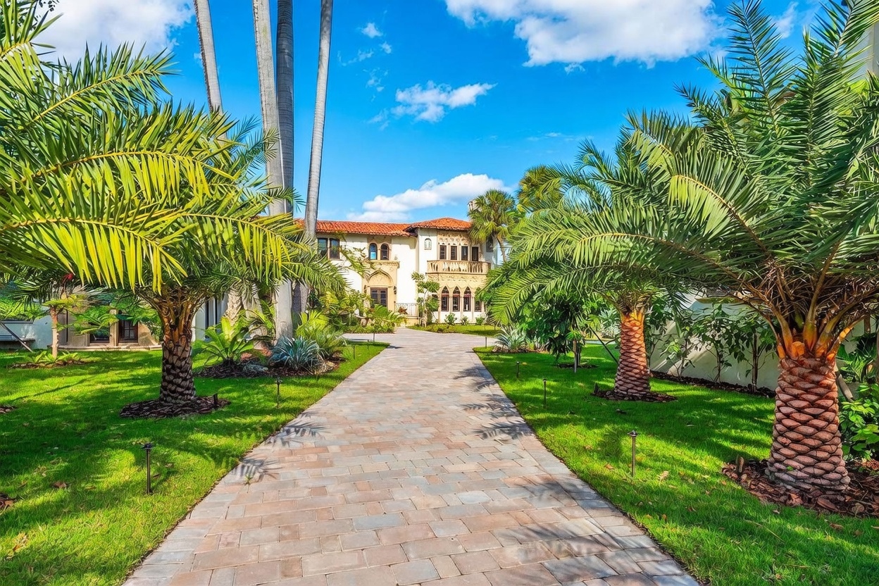 This Mediterranean-style estate features a grand, light-colored stucco facade with intricate architectural detailing and a classic red-tiled roof. A long, paved driveway leads directly to the main entrance, flanked by lush green lawns and mature palm trees that frame the view. The perspective is a centered, eye-level shot that emphasizes the symmetry and stately curb appeal of the property.