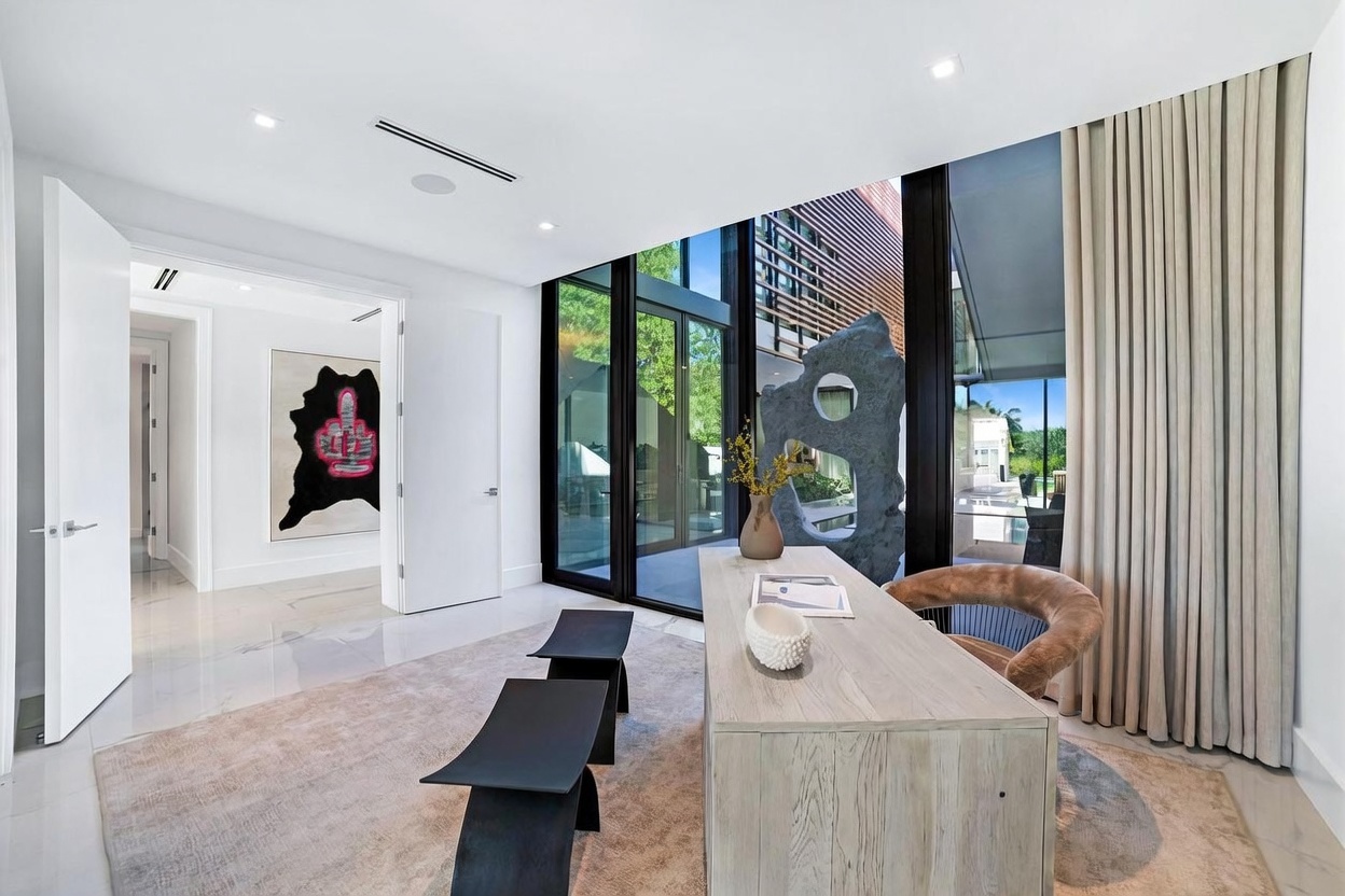 This modern home office features a light wood desk positioned on a large area rug, complemented by a curved chair with a textured seat. The room is defined by expansive floor-to-ceiling windows that provide a view of an outdoor sculpture and pool area, while a piece of abstract art hangs on the wall near the hallway entrance. The space is bright and minimalist, with polished light-colored flooring and clean white walls creating a sophisticated, airy atmosphere.
