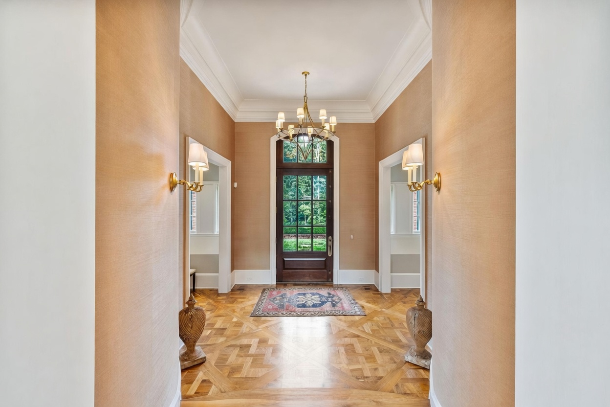 This elegant foyer features intricate patterned wood flooring and warm, textured wall coverings that create a sophisticated entrance. A classic chandelier hangs from the high ceiling, flanked by two wall-mounted sconces that illuminate the path toward a tall, multi-paned glass front door. The perspective is centered and symmetrical, offering a clear view through the entryway to the lush greenery outside.